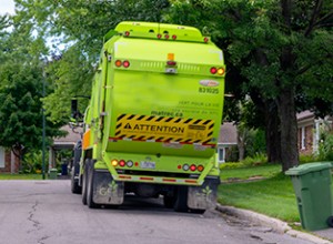 Camion à ordures vert en bordure de rue avec un bac vert à droite.