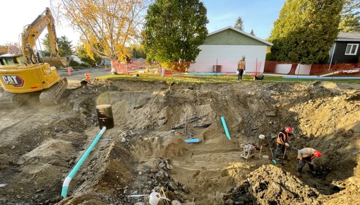 Rue creusée par une pelle mécanique jaune afin de remplacer des tuyaux d'aqueduc municipaux. Deux travailleurs sont à droite de la photo. On voit 2 maisons au loin.