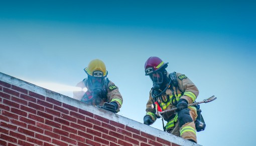 Deux pompiers sur le toit d'un bâtiment en brique, vus en plongée.